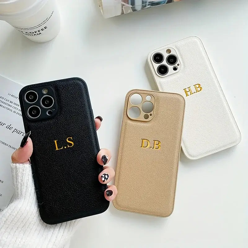 Three personalized leather iPhone cases in black, beige, and white with initials L.S, D.B, and H.B, displayed on a table.